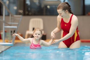 DLRG Rettungsschwimmerin führt ein kleines Mädchen an der Hand ins Hallenbad-Becken.