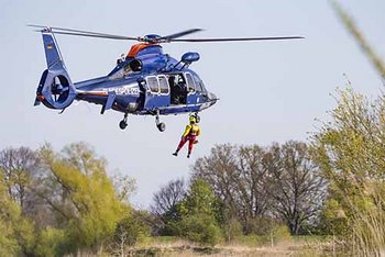 Luftretter in rot-gelber Einsatzkleidung hängt seitlich aus blauem Hubschrauber