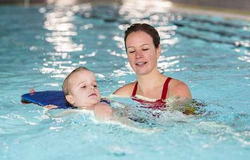 Kind übt mit Schwimmbrett im Nacken und Ausbilderin unterstützt