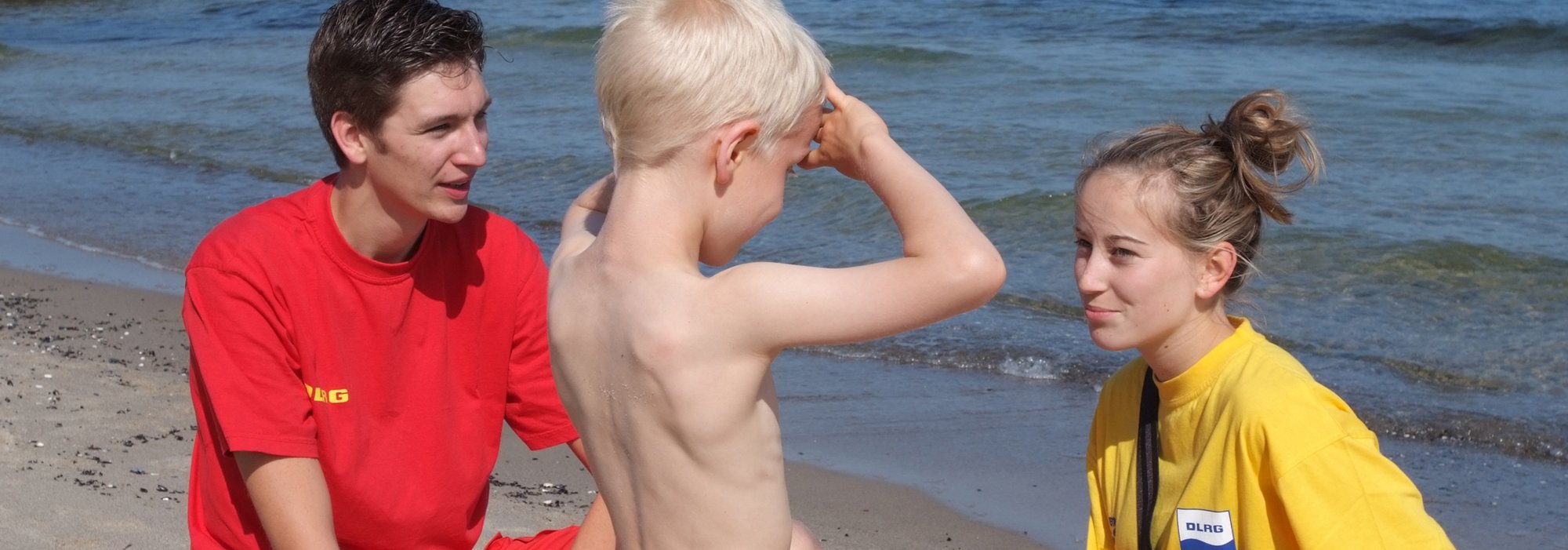 Zwei Rettungsschwimmer unterhalten sich mit einem Kind am Strand.