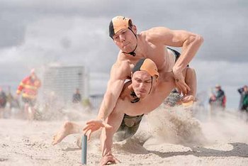 Zwei Rettungssportler greifen im Sprung nach Stab im Strand