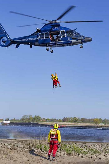 Luftretter in rot-gelber Einsatzkleidung seilen sich von blauem Helikopter ab über See