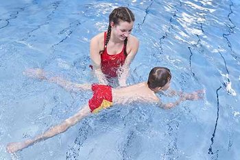 Kind übt Schwimmen im Hallenbad mit ganzem Körper, unterstützt von Ausbilderin
