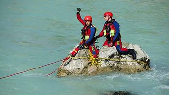 Zwei Strömungsretter mit roten Helmen und rot-blauer Einsatzkleidung sitzen auf Felsen in fließendem Gewässer mit gespannter Rettungsleine